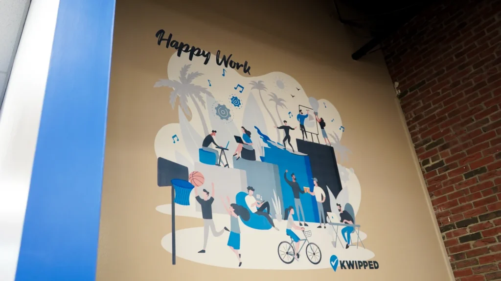 A Happy Work illustration of people at desks printed on an office wall beside exposed brick