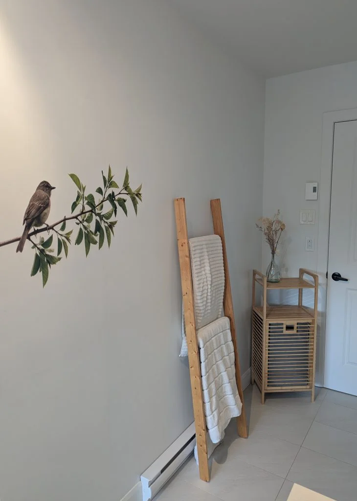 A bird on a leafy branch printed on a white wall beside a towel ladder