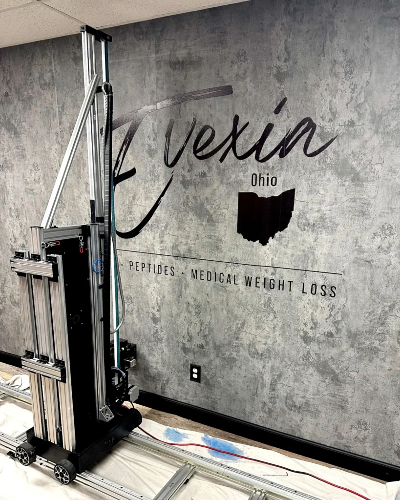 Script logo lettering printed on a textured gray wall, with the vertical printer in front