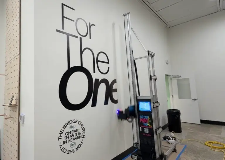Wall printing machine mid-print on a white church wall with black For The One lettering and The Bridge Church seal
