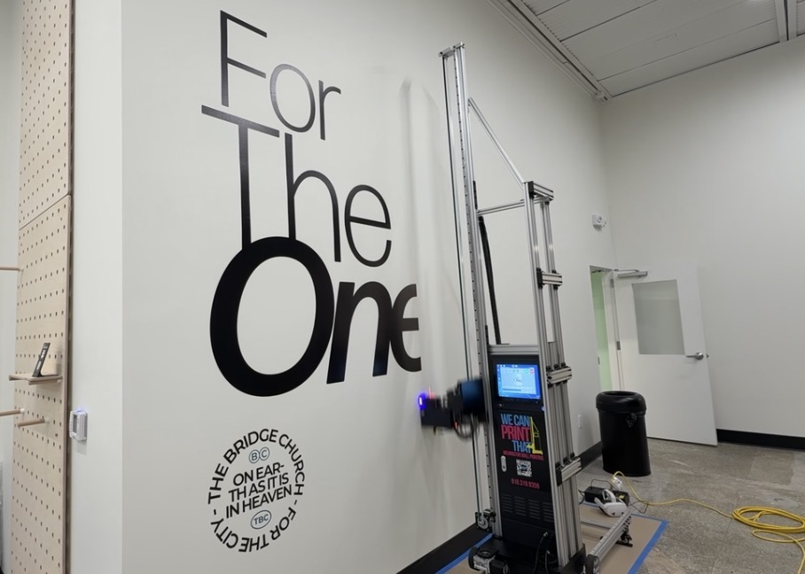 Wall printing machine mid-print on a white church wall with black For The One lettering and The Bridge Church seal