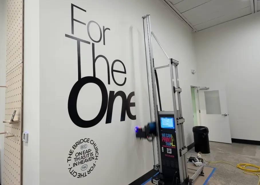 Wall printing machine mid-print on a white church wall with black For The One lettering and The Bridge Church seal